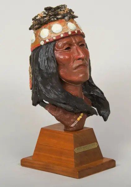 Clay bust of a Native American chief with a beaded orange headband and black feathered headdress on a wooden pedestal; plaque reads 'White Mountain Chief'.