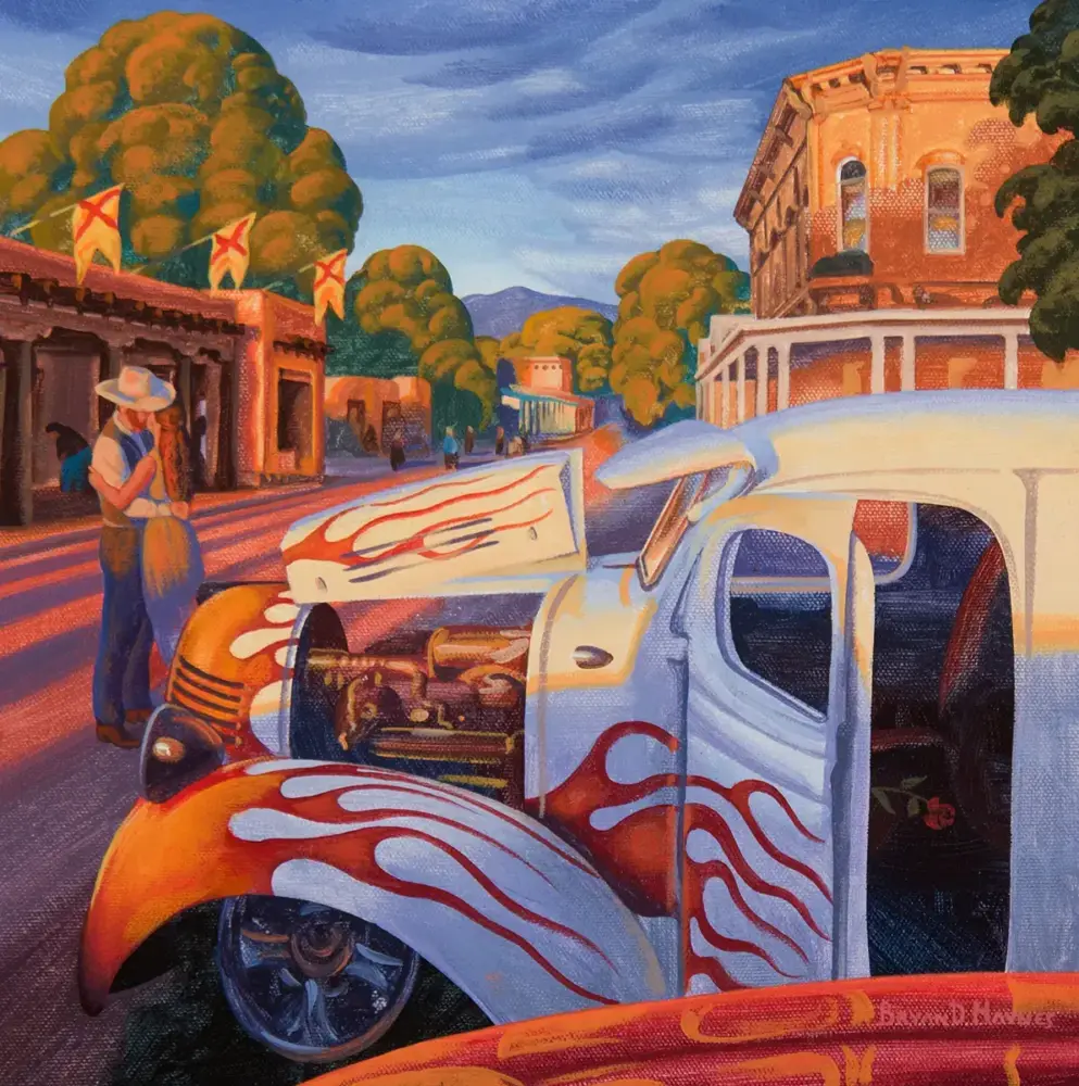 Colorful vintage hot rod with orange flames parked on a sunlit street, with a man in a hat near buildings and trees.