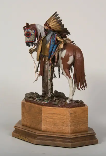 Mixed-media sculpture of a Native American man with a feathered headdress guiding a pinto horse on a wooden pedestal base.