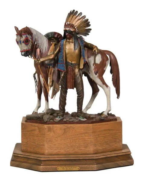 Life-like sculpture of a Native American man with a tall feather headdress standing beside a brown-and-white horse on a wooden pedestal, holding the horse's reins