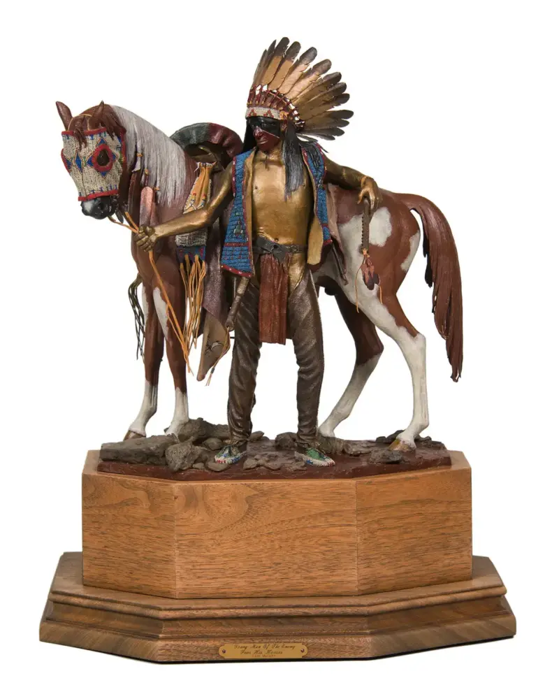 Life-like sculpture of a Native American man with a tall feather headdress standing beside a brown-and-white horse on a wooden pedestal, holding the horse's reins