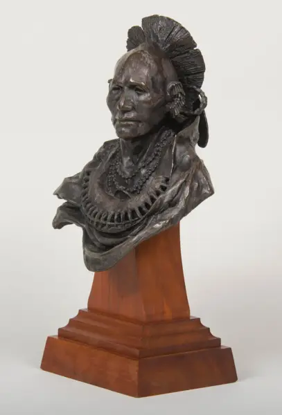 Bronze bust of a Native American man with a feathered headdress and beaded necklace, mounted on a wooden pedestal.