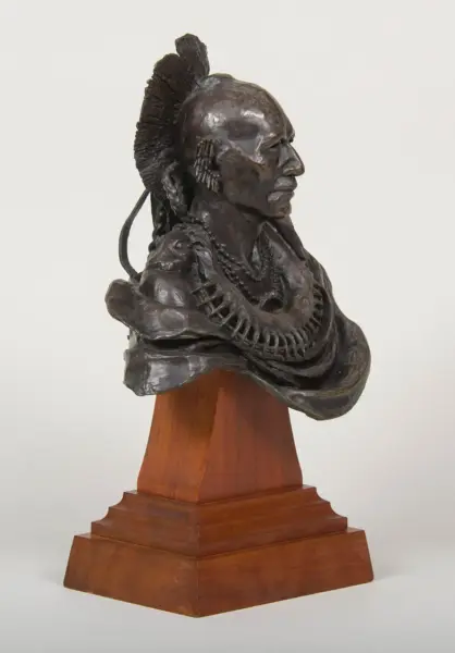 Bronze bust sculpture of a Native American man with a tall mohawk and bead necklace on a wooden pedestal.