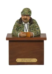 Carved figurine of a camo-clad hunter at a wooden podium, hands on a wallet; front plaque with a hunting joke.