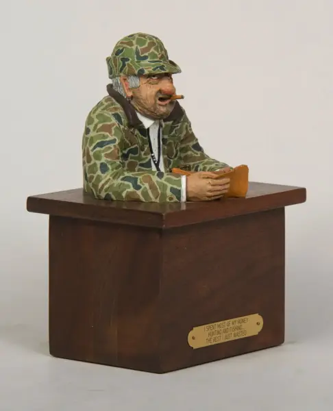 Humorous carved wooden desk figurine of an old hunter in camouflage, seated at a lectern with a cigar and a folded bill.