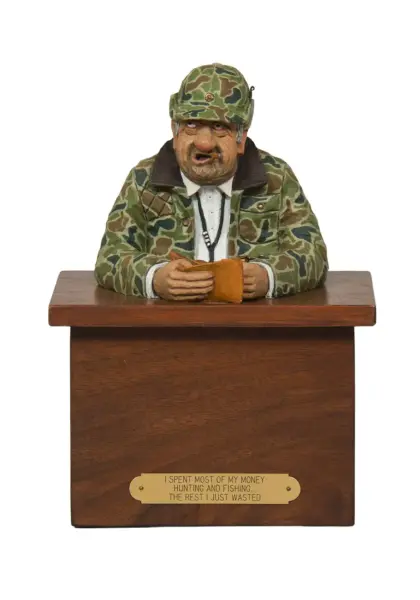 Carved figurine of a camo-clad hunter at a wooden podium, hands on a wallet; front plaque with a hunting joke.