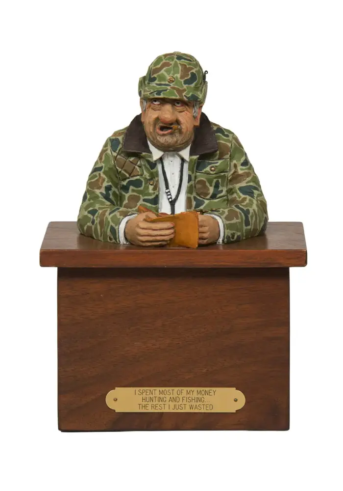 Carved figurine of a camo-clad hunter at a wooden podium, hands on a wallet; front plaque with a hunting joke.