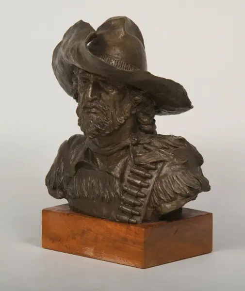 Bronze bust sculpture of a bearded cowboy in a wide-brim hat with a bandolier across his chest, mounted on a wooden pedestal.