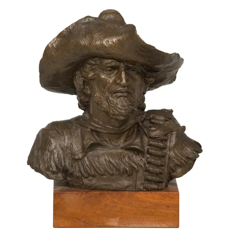 Bronze bust of a rugged cowboy with a wide-brimmed hat and beard, mounted on a wooden plinth.