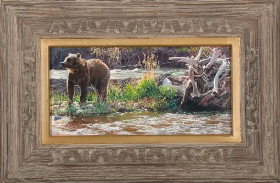 Framed painting of a brown bear standing by a river with grasses and driftwood nearby.