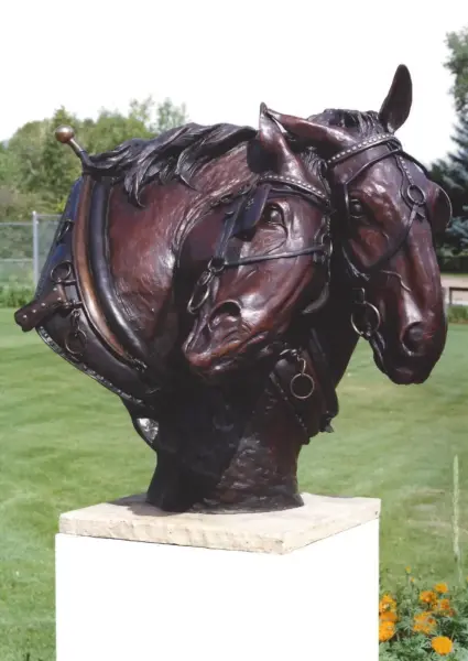 Bronze sculpture of two horse heads with bridles, mounted on a white pedestal in a grassy park